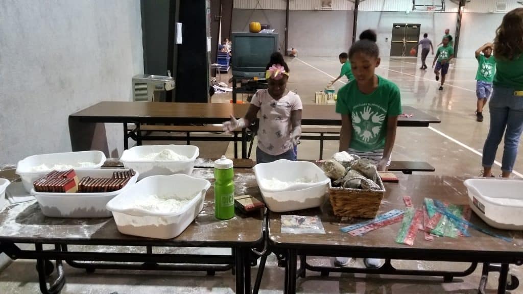 Kids standing at tables with tubs of flour, rocks, and rulers during a hands-on science activity in a gym.