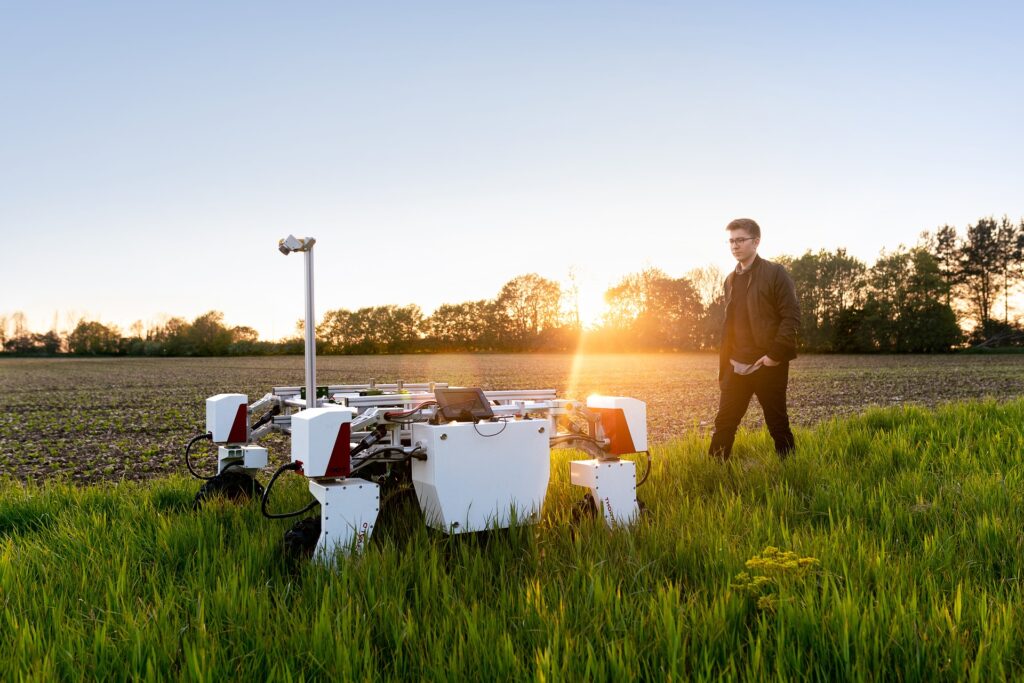 Engineer field‑testing a robotic platform in a crop field at sunset, showing how small R&D labs trial new technology in real‑world conditions before rollout.