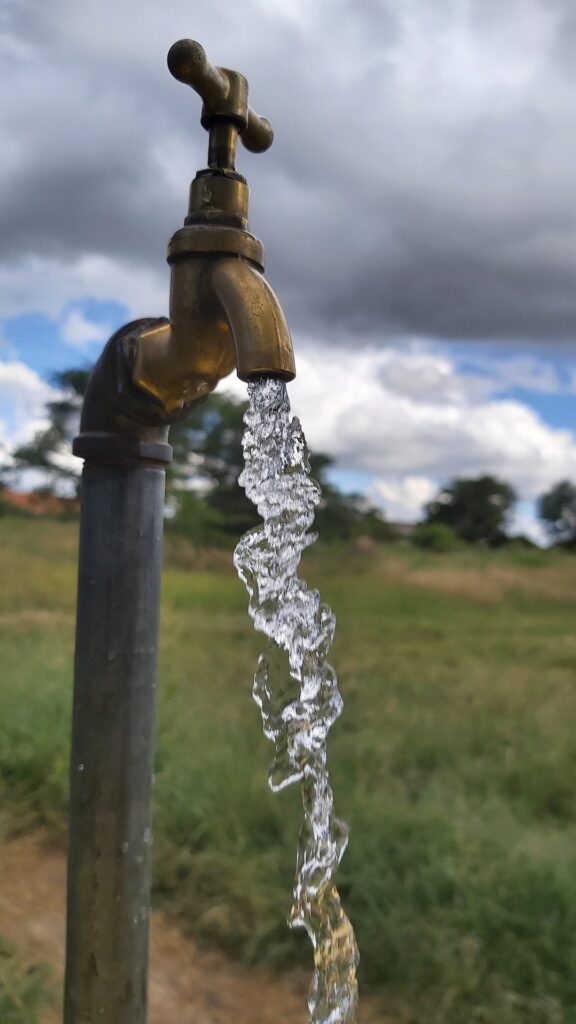 Brass outdoor water tap with a continuous stream of clear water against a cloudy sky and green field, symbolizing rural access to safe drinking water.