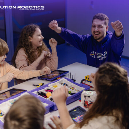 Children and instructor celebrating around a table during an in-person Revolution Robotics class, with robotics kits and iPads spread out in front of them.