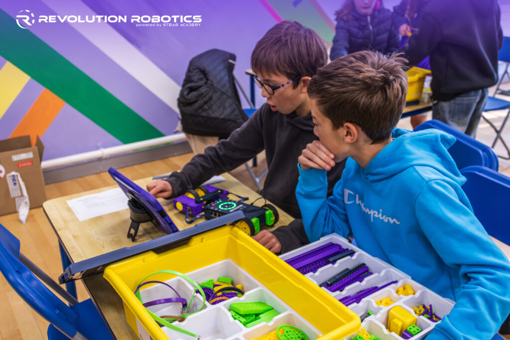 Two boys in a colorful classroom work together at a table, using a tablet and a wheeled robot kit with bins of bright plastic parts, as they iteratively build and test their design.
