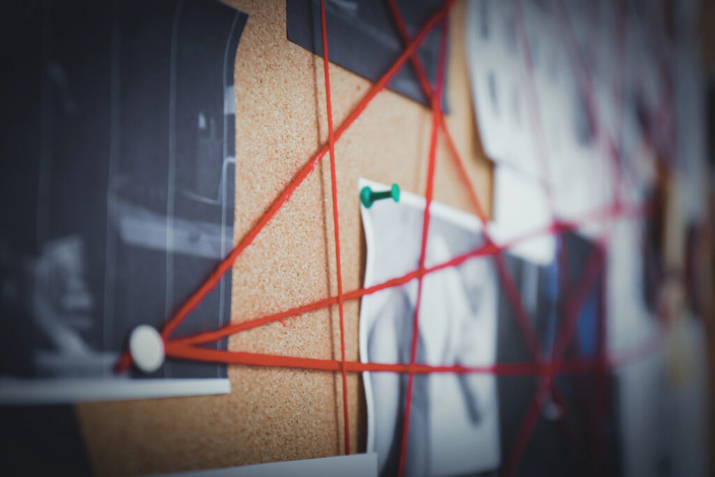 Close-up of a cork investigation board with photographs connected by red string and secured with pushpins.