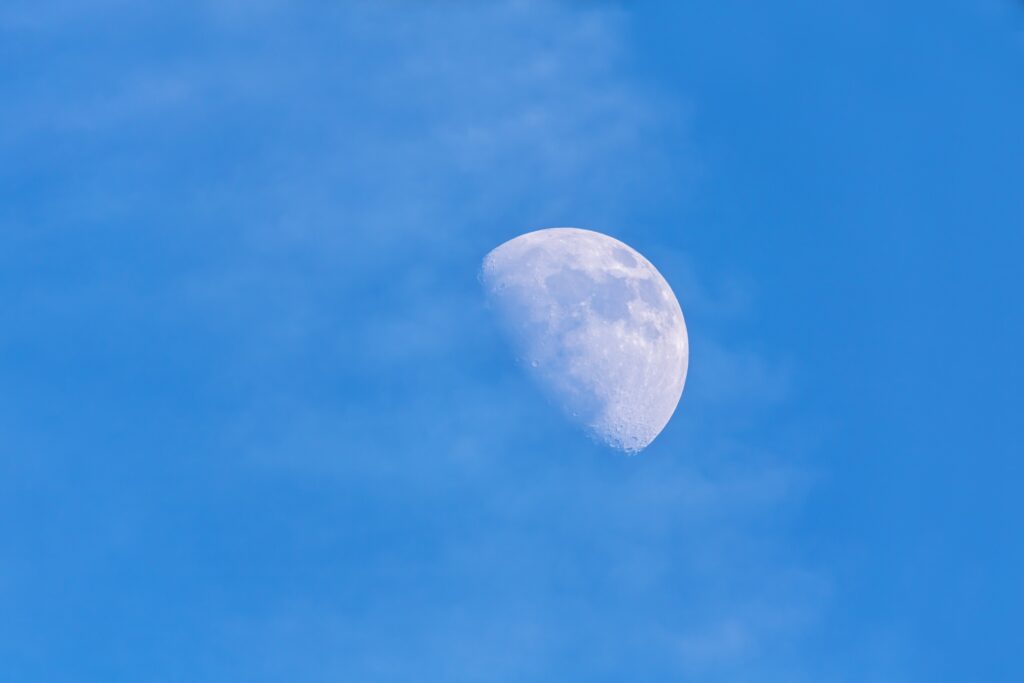 Partially illuminated Moon against a blue daytime sky, with visible craters and darker patches across its sunlit surface.